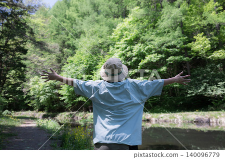 A woman takes a deep breath and spreads her arms while enjoying a forest bath; a faceless image showing her back as she enjoys the outdoors and summer camping. 140096779
