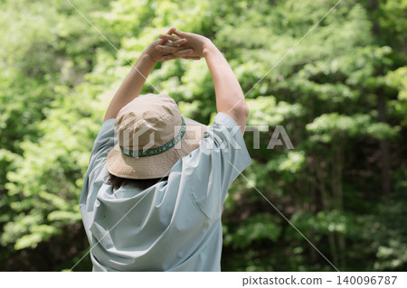 A woman takes a deep breath and spreads her arms while enjoying a forest bath; a faceless image showing her back as she enjoys the outdoors and summer camping. A woman takes a deep breath and spreads her arms while enjoying a forest bath; a faceless image showing her back as she enjoys the outdoors and summer camping. 140096787