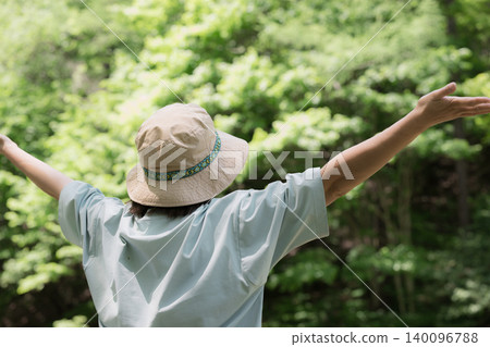 A woman takes a deep breath and spreads her arms while enjoying a forest bath; a faceless image showing her back as she enjoys the outdoors and summer camping. A woman takes a deep breath and spreads her arms while enjoying a forest bath; a faceless image showing her back as she enjoys the outdoors and summer camping. 140096788