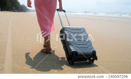 Close rear view of young woman in pink dress rolling a grey suitcase along sandy beach with sea waves nearby. Summer vacation concept 140097206