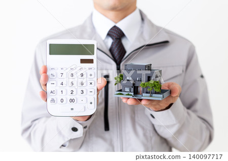 A middle-aged businessman in gray work clothes, holding a model house and a calculator, against a white background. A middle-aged businessman in gray work clothes, holding a model house and a calculator, against a white background. 140097717