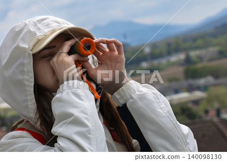 Young woman looking through a small orange telescope at a river landscape, highlighting lakeside and mountain tourism. Young woman looking through a small orange telescope at a river landscape, highlighting lakeside and mountain tourism. 140098130
