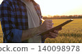 Close-up of male farmer holding pen and clipboard, writing notes during crop inspection in wheat field at sunset. Concept of agricultural documentation, crop control, and farm work. 140098978
