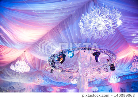 Luxurious event ceiling with crystal chandeliers, draped fabric, and pink and purple lighting Luxurious event ceiling with crystal chandeliers, draped fabric, and pink and purple lighting 140099068