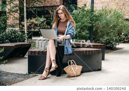 Woman uses laptop while sitting on wooden bench outdoors. Work-from-home alternatives, outdoor remote work, nomadic work lifestyle. 140100248