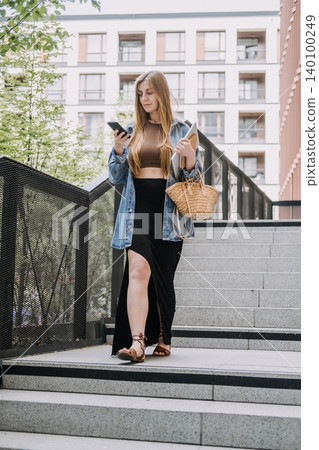 Young woman stands on city stairs looking at smartphone while holding laptop. On the fly decision making, active professional lifestyle, business mobility trends. Young woman stands on city stairs looking at smartphone while holding laptop. On the fly decision making, active professional lifestyle, business mobility trends. 140100249