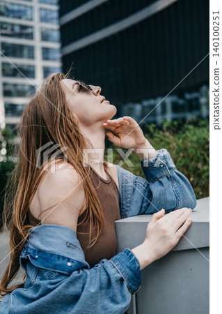 Woman wearing sunglasses tilts head up while leaning against outdoor structure in corporate district. Stress management metropolitan, taking a mindful break, personal resilience city dweller. Woman wearing sunglasses tilts head up while leaning against outdoor structure in corporate district. Stress management metropolitan, taking a mindful break, personal resilience city dweller. 140100251