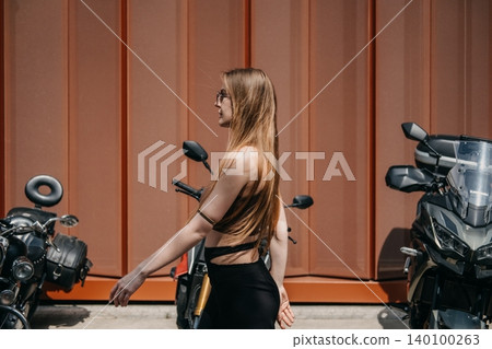 Woman walks past parked motorcycles in front of textured wall. Independent woman traveling, modern urban lifestyle, confident female walking. 140100263