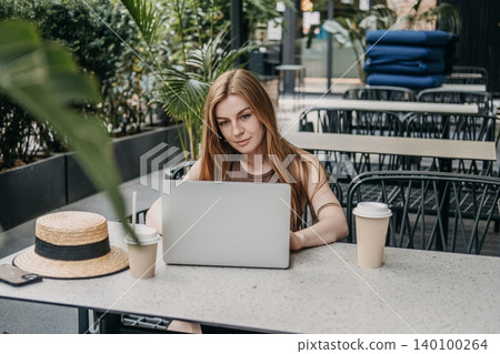 Woman sits at outdoor cafe table using laptop computer. Digital nomad lifestyle, remote work anywhere, flexible working hours. Woman sits at outdoor cafe table using laptop computer. Digital nomad lifestyle, remote work anywhere, flexible working hours. 140100264