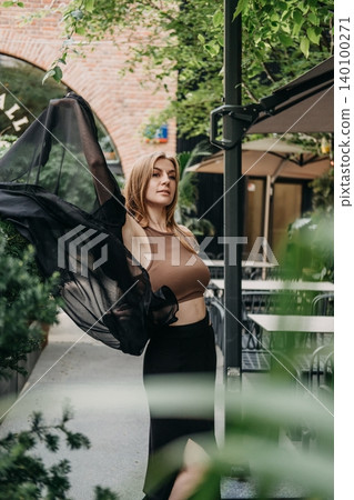 Woman holds up sheer black fabric while posing on outdoor cafe patio. Urban fashion portrait, contemporary street style, modern female look. Woman holds up sheer black fabric while posing on outdoor cafe patio. Urban fashion portrait, contemporary street style, modern female look. 140100271