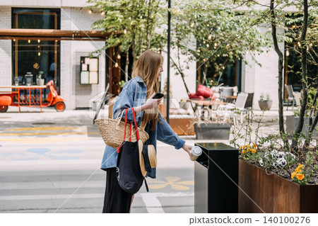 Woman drops paper cup into black outdoor public trash bin. Disposing takeaway cup, throwing garbage in bin, commercial public trash. Woman drops paper cup into black outdoor public trash bin. Disposing takeaway cup, throwing garbage in bin, commercial public trash. 140100276
