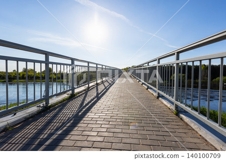 Sunlit pedestrian bridge crossing a calm river, featuring a paved walkway Sunlit pedestrian bridge crossing a calm river, featuring a paved walkway 140100709