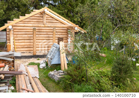 A partially built log cabin sits amid a green garden with stacked lumber 140100710