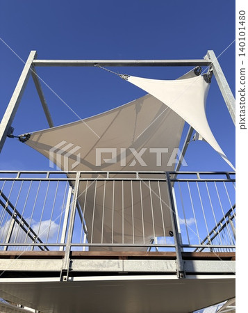 a modern tensile fabric structure, likely a large shade canopy or sail, set against a vibrant blue sky a modern tensile fabric structure, likely a large shade canopy or sail, set against a vibrant blue sky 140101480