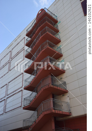 A modern building features a prominent red fire escape staircase attached to its exterior wall. The staircase has multiple levels and is surrounded by a light-colored facade under a clear blue sky 140102621