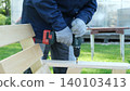 Using a cordless drill driver to drive screws into a wooden chair. The craftsman holds the tool securely with protective gloves. Traditional carpentry. Safety precautions. DIY repair. Copy space 140103413
