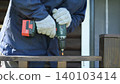 Using a cordless cordless drill driver to drive screws into a wooden porch railing. The craftsman holds the tool securely with protective gloves. Traditional carpentry. Safety precautions. DIY repair 140103414