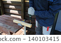 Using a cordless drill driver to drive screws into a wooden board on an outdoor workbench. Gloved hands hold the tool securely. The board is held in place with a clamp. Safety precautions. DIY repair 140103415