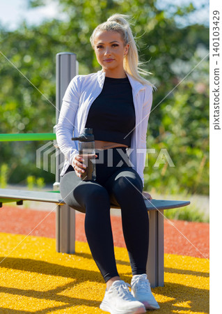 Woman resting outdoors in activewear holding water bottle on a sunny day 140103429
