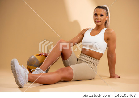 Fit woman in white sports bra and beige leggings sitting with medicine ball on floor Fit woman in white sports bra and beige leggings sitting with medicine ball on floor 140103468