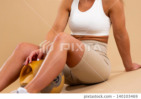Athletic woman in white sports bra and beige leggings sitting with yellow medicine ball on floor 140103469