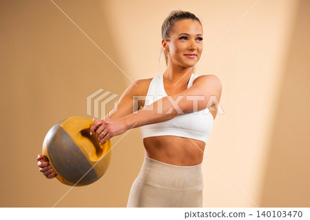 Fit woman in white workout gear holding a yellow and gray kettlebell with a confident smile 140103470