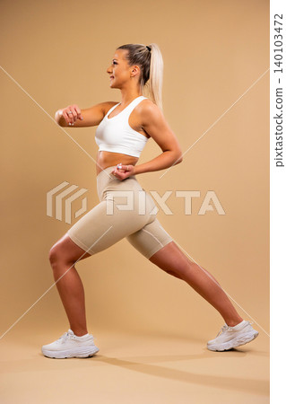 Athletic woman in white sports bra and beige leggings performing lunges in studio setting Athletic woman in white sports bra and beige leggings performing lunges in studio setting 140103472