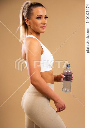 Fit woman in white sports bra and beige leggings holding water bottle standing in studio Fit woman in white sports bra and beige leggings holding water bottle standing in studio 140103474