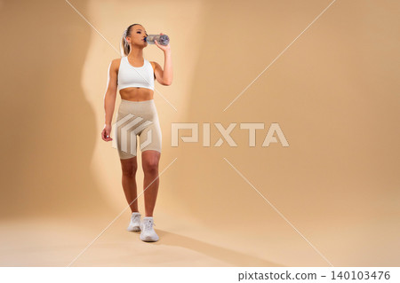 Fit athletic woman in white sports bra and beige leggings drinking water from bottle during workout session 140103476