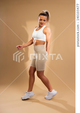 Fit young woman in white sports bra and beige leggings walking confidently in studio setting Fit young woman in white sports bra and beige leggings walking confidently in studio setting 140103477