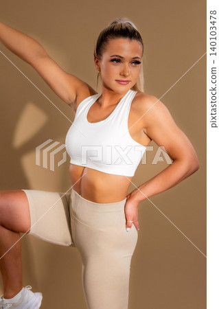 Fit young woman in white sports bra and beige leggings posing confidently in fitness wear Fit young woman in white sports bra and beige leggings posing confidently in fitness wear 140103478