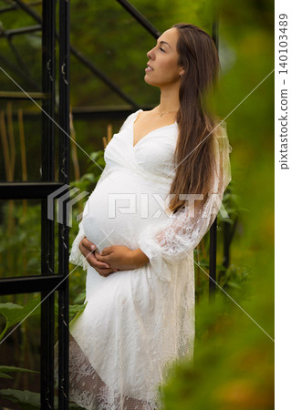 Pregnant woman in white dress reflecting in a lush green garden 140103489