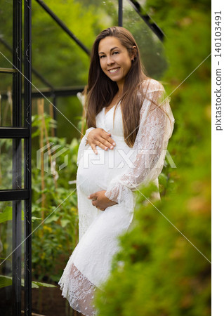 Smiling pregnant woman in lace dress surrounded by greenery and natural light 140103491