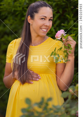 Pregnant woman in a yellow dress holding a pink rose, smiling outdoors 140103493