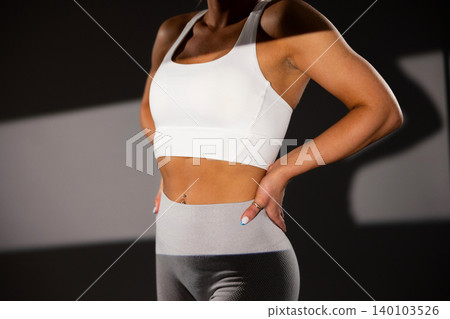 Fit woman in white sports bra and gray leggings standing confidently in gym setting Fit woman in white sports bra and gray leggings standing confidently in gym setting 140103526