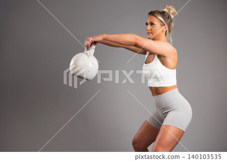 Athletic woman performing kettlebell swing in white workout gear on gray background Athletic woman performing kettlebell swing in white workout gear on gray background 140103535
