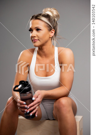 Fit young woman in white workout tank holding water bottle while sitting on wooden box Fit young woman in white workout tank holding water bottle while sitting on wooden box 140103542