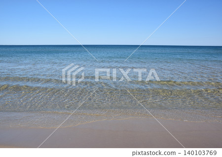 A serene beach landscape featuring clear blue water meeting a soft sandy shore, with gentle waves creating a calming effect under a clear sky, ideal for relaxation and leisure 140103769