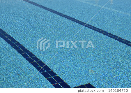 A clear swimming pool with blue water, featuring lane markings in dark blue. The surface reflects light, creating a shimmering effect across the water's surface A clear swimming pool with blue water, featuring lane markings in dark blue. The surface reflects light, creating a shimmering effect across the water's surface 140104750