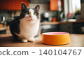 Black and white cat sits in the kitchen near an orange bowl. Bright green eyes glow under warm light. 140104767