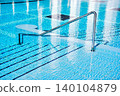 A swimming pool features a stainless steel handrail leading to tiled steps submerged in clear water, with reflections of the surrounding area visible on the water's surface 140104879