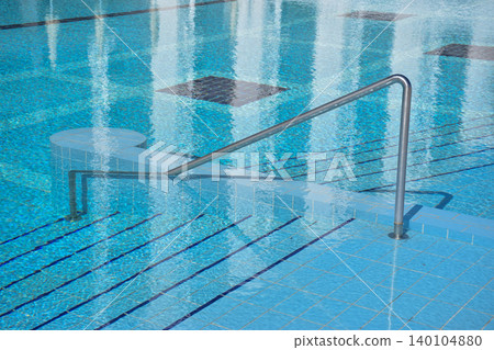 A swimming pool features a stainless steel handrail leading to tiled steps submerged in clear water, with reflections of the surrounding area visible on the water's surface A swimming pool features a stainless steel handrail leading to tiled steps submerged in clear water, with reflections of the surrounding area visible on the water's surface 140104880