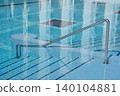 A swimming pool features a stainless steel handrail leading to tiled steps submerged in clear water, with reflections of the surrounding area visible on the water's surface 140104881