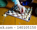 Two children playing chess on a wooden floor. One child has light skin and wears colorful socks. The other child has dark skin and is focused on the game. 140105150