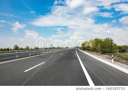 Wide highway with clear blue sky, guardrail, roadside vegetation Wide highway with clear blue sky, guardrail, roadside vegetation 140105733