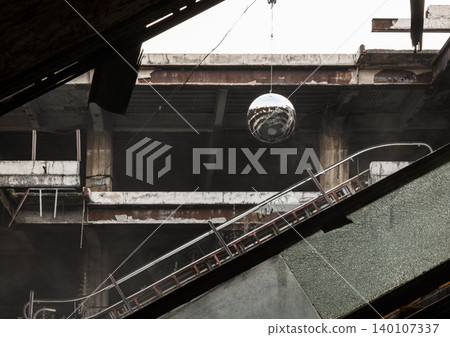 Concrete decay meets modern art in a silent, abandoned space. Neglect is disrupted by a sudden glimmer of life. Weathered concrete and rusted girders frame the hollow interior. A broken escalator cuts 140107337