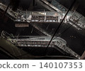 Fractured, multi-tiered escalators cut diagonally across a cavernous void, their rusted undercarriages exposing grid-like steel ribbing. Deep shadows swallow the background, sharply contrasting with 140107353