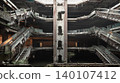 The skeletal ruins of an abandoned shopping mall rise symmetrically around a central shaft, their rusted escalators frozen over a dark, flooded floor. Patches of wild green foliage drape softly across 140107412