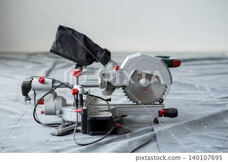 Electric miter saw with dust bag on protective plastic floor covering during home renovation works Electric miter saw with dust bag on protective plastic floor covering during home renovation works 140107695