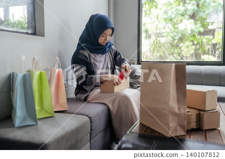 Woman in hijab preparing a gift at home with shopping bags and cardboard boxes near a window Woman in hijab preparing a gift at home with shopping bags and cardboard boxes near a window 140107812
